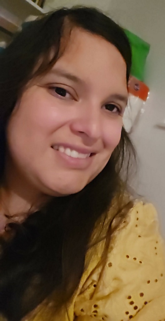 Photo of Dominique Sapien: Close-up portrait of a smiling woman with long dark hair wearing a yellow top, photographed indoors with a warm, friendly expression.