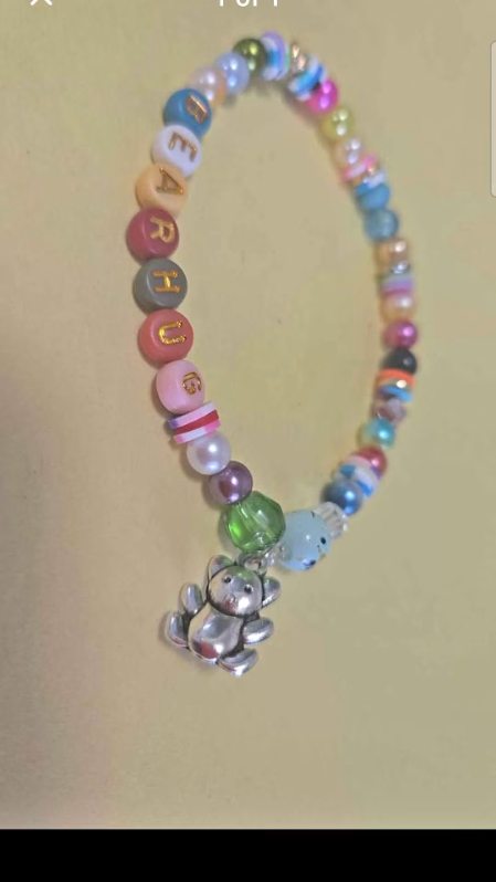 Site Logo: Colorful beaded bracelet with pastel and bright beads, letter beads, and a dangling silver bear charm on a beige background.